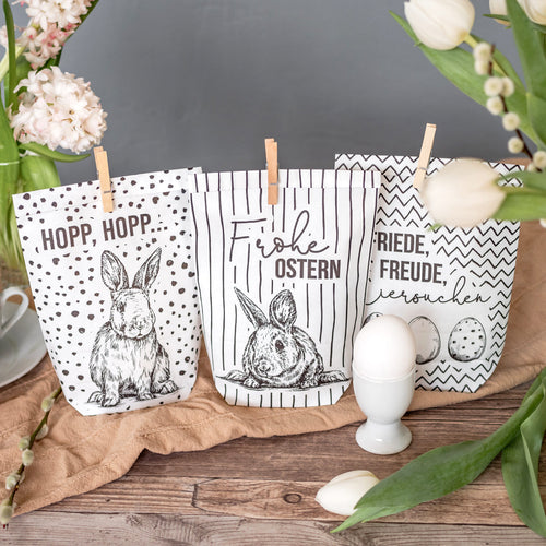 Drei schwarz weiße Wundertüten mit verschiedenen Musterungen gezackt, gepunktet, gestreift. Bedruckt mit den Schriften "Hopp, Hopp", "Friede Freude, Eierkuchen" und "Frohe Ostern" und verschiedenen Hasen auf dem Tisch