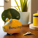 Gelbes Sparschwein aus Keramik mit grauer Aufschrift: Benzingeld, angerichtet auf einem Holztisch mit Pflanzen im Hintergund und schlüssel im Vordergrund