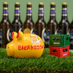 Keramik Sparschwein "Bierkasse" gelb und rot, fotografiert auf Kunstrasen mit Bierkästen und Bierflaschen im Hintergund, im Sparschwein steckt ein goldene Münze