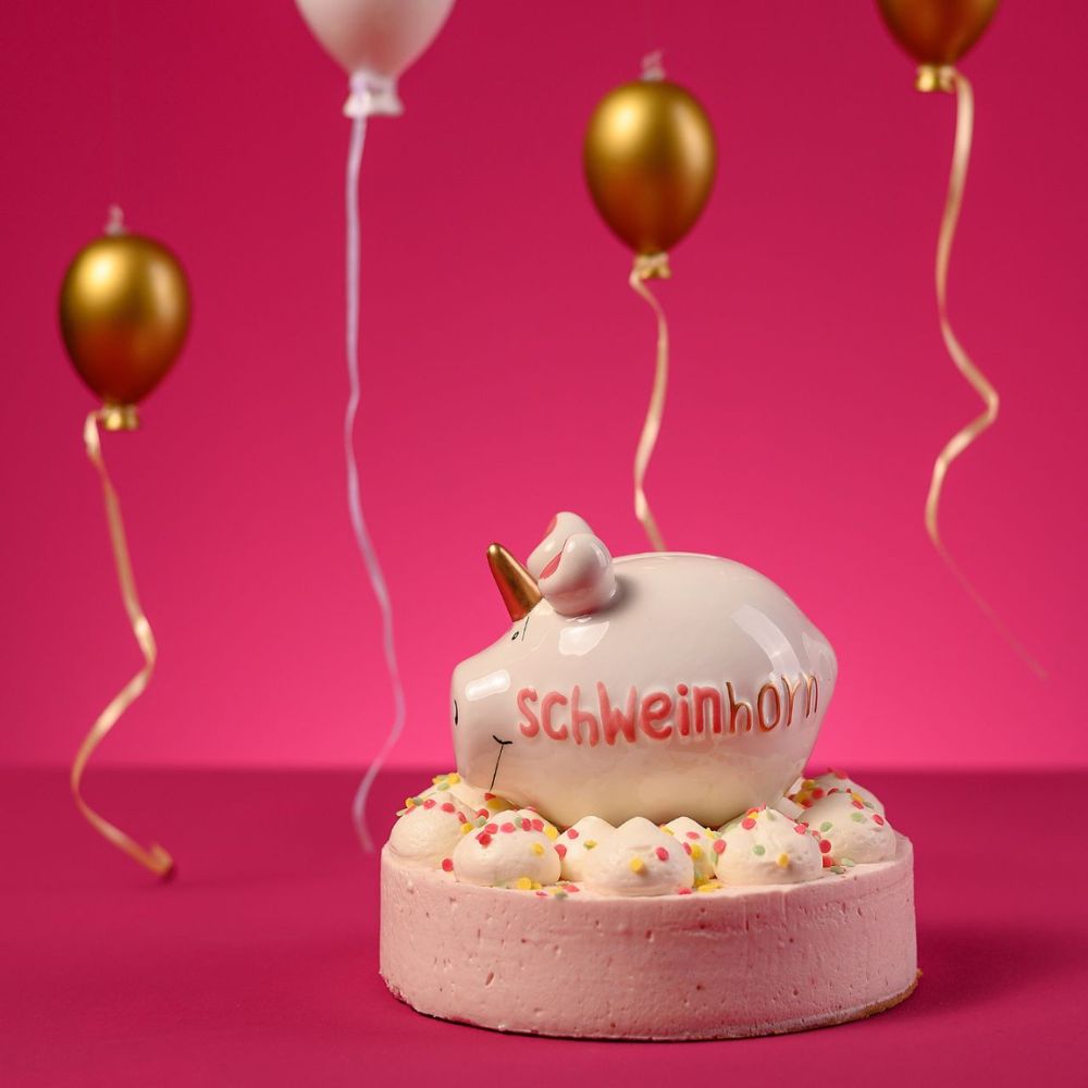 Keramik Spardose "Schweinhorn" in weiß rosa und Gold, fotografiert vor pinkem Hintergrund mit rosanen und goldenen Luftballons, sitzend in einer rosanen Torte mit bunten Streuseln