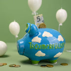 Großes Sparschwein aus Keramik, tiefblau mit weißes Wolken, Grüne Details und grüner Schriftzug "Traumerfüller", Gesichtszüge des Sparschweins in schwarz gemalt, fotografiert vor grünem hintergund mit weißen Luftballons, weiße Noppen, goldene Münzen