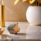 Weißes Sparschwein mit schwarzen Füßen und Ohren und der Schrift "Luxus Schwein" Die Schrift ist umgeben mit goldenen ovalen Formen. Das Schweinchen steht auf einem marmor Thresen mit goldenen Deko-Elementen