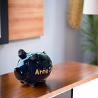 Schwarzes Sparschwein aus Keramik, graue Details (Schweinohren und Füße) Noppen an der Unterseite , Gesichtzüge in weiß, weiße große Aufschrift: Arme Sau, Sparschwein fotografiert auf Holztisch