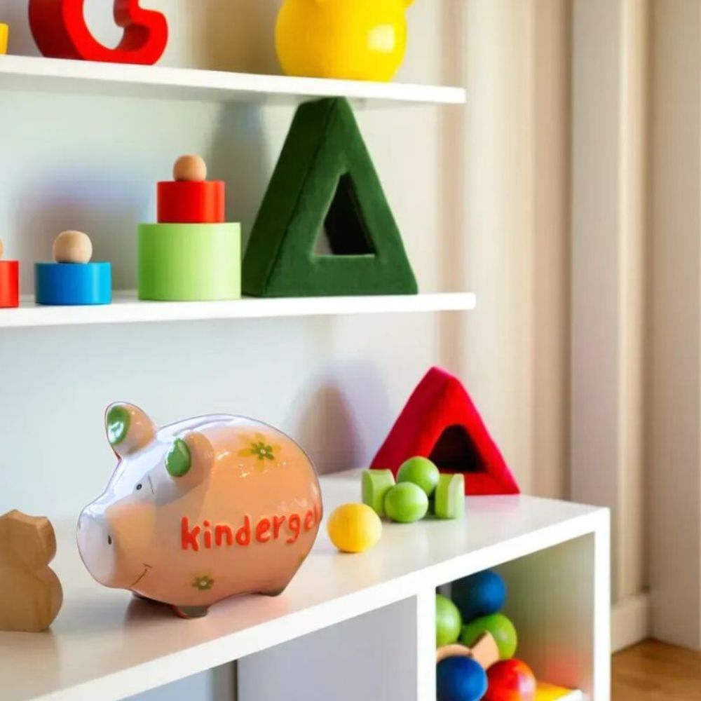 Sparschwein aus Keramik in Weiß, grüne Details (Schweineohren und Füße), Grüne Blümchen, orange-roter Schriftzug "kindergeld" auf der Seite des Sparschweins, Gesichtszüge gemalt in Schwarz, angerichtet in einem Kinderzimmer mit bunten Spielzeugen