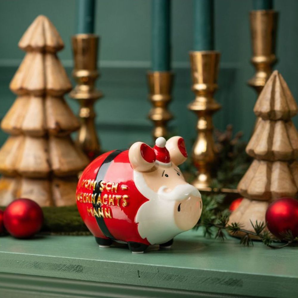 Sparschwein mit weißem Bart und rotem Weihnachtsmann Anzug und einer Goldenen Schrift "Vom Sch-Weihnachtsmann" auf einem grünen Thresen mit Weihnachtsdeko im Hintergrund