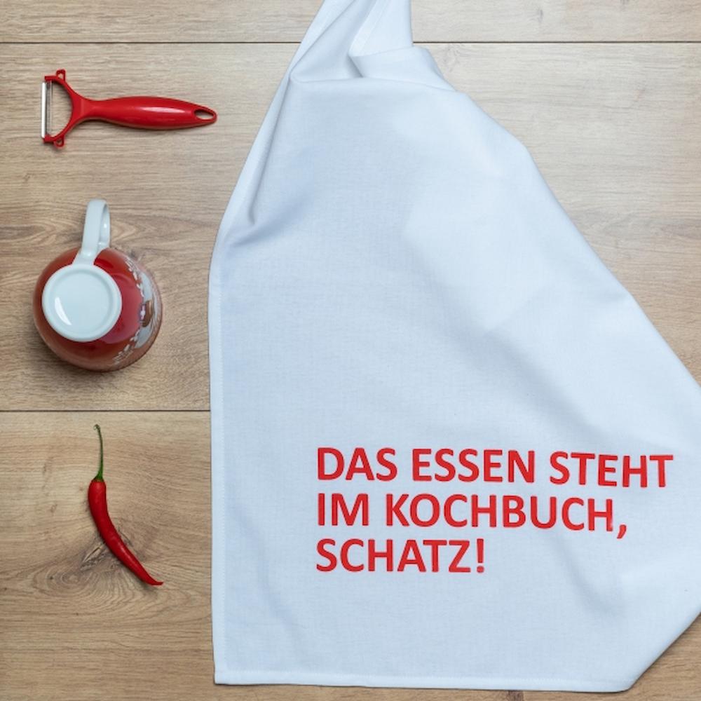Weißes Geschirrtuch mit einer Roten Schrift "DAS ESSEN STEHT IM KOCHBUCH, SCHATZ!" auf einem Tisch mit Chili, einer Tasse und einem Roten Kartoffelschäler
