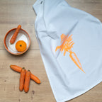 Weißes Geschirrtuch mit einem orangenen Audruck einer Karotte ausgebreitet auf einem Tisch auf dem eine Schale steht und Möhren liegen 