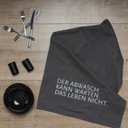 Graues Baumwoll-Geschirrtuch mit einer weißen aufgedruckten Schrift "DER ABWASCH KANN WARTEN. DAS LEBEN NICHT." auf einem Tisch mit Besteck und einer Schüssel