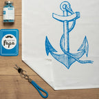 Das Baumwoll Geschirrtuch mit einem aufgedruckten blauen Anker liegt auf einem Tisch umgeben mit blauem Dekor