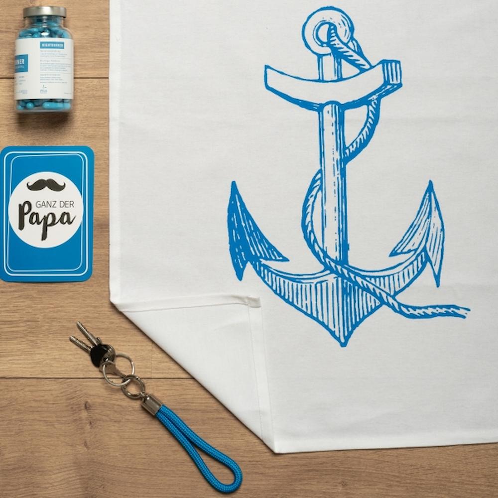 Das Baumwoll Geschirrtuch mit einem aufgedruckten blauen Anker liegt auf einem Tisch umgeben mit blauem Dekor