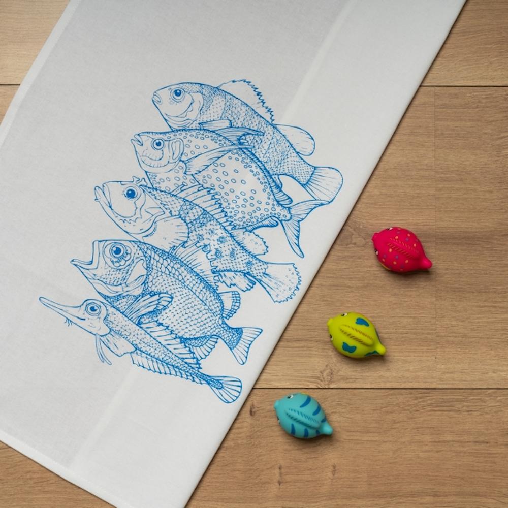 Weißes Geschirrtuch mit einem blauen Aufdruck von 5 Fischarten auf einem Tisch ausgebreitet mit drei Deko Fischen