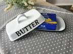 weiße Butterdose mit der schwarzen Schrift "BUTTER", geöffnet mit enthaltener Butter auf einem Tisch