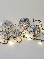 Auf einem weißen Boden liegende Weinachtskugeln mit einem Delfterblauen Keramik Design, aus Papier zusammengesteckt und einer leutchtenden Lichterkette