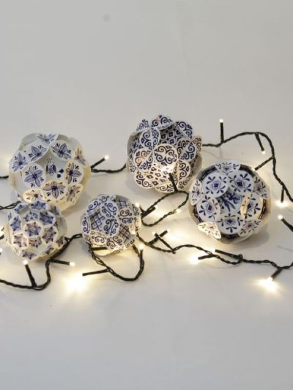 Auf einem weißen Boden liegende Weinachtskugeln mit einem Delfterblauen Keramik Design, aus Papier zusammengesteckt und einer leutchtenden Lichterkette