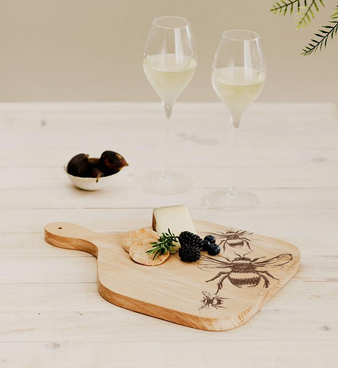 Servierpaddel aus Eiche mit eingebranntem Bienenmuster liegt auf einem Tisch mit zwei Gläsern Champagner