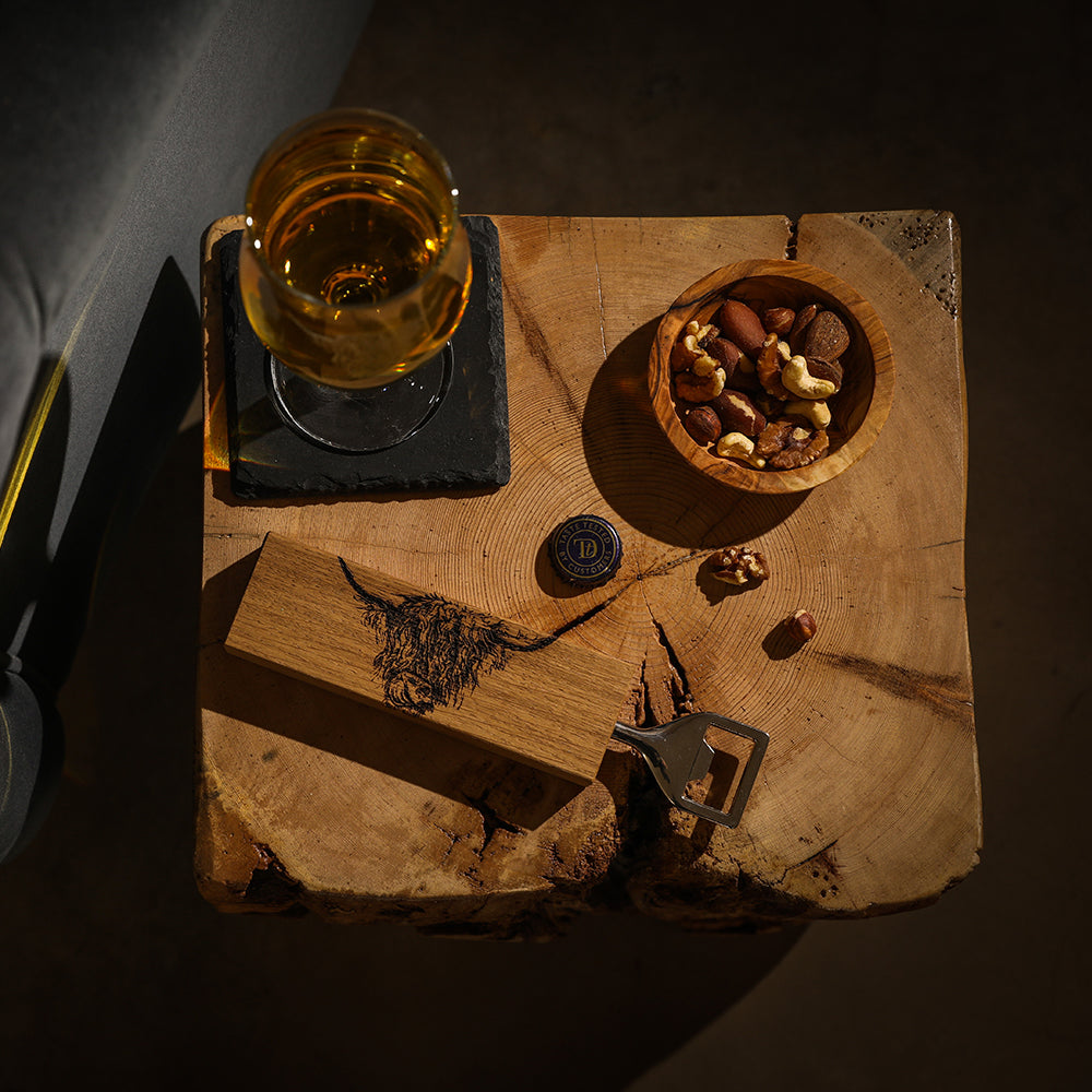 Ein Flaschenöffner mit einem rechteckigen Eichenholzgriff, auf dem ein Hochlandrind-Motiv eingebrannt ist dargestellt mit einer Nuss-Schale und einem Glas Whisky