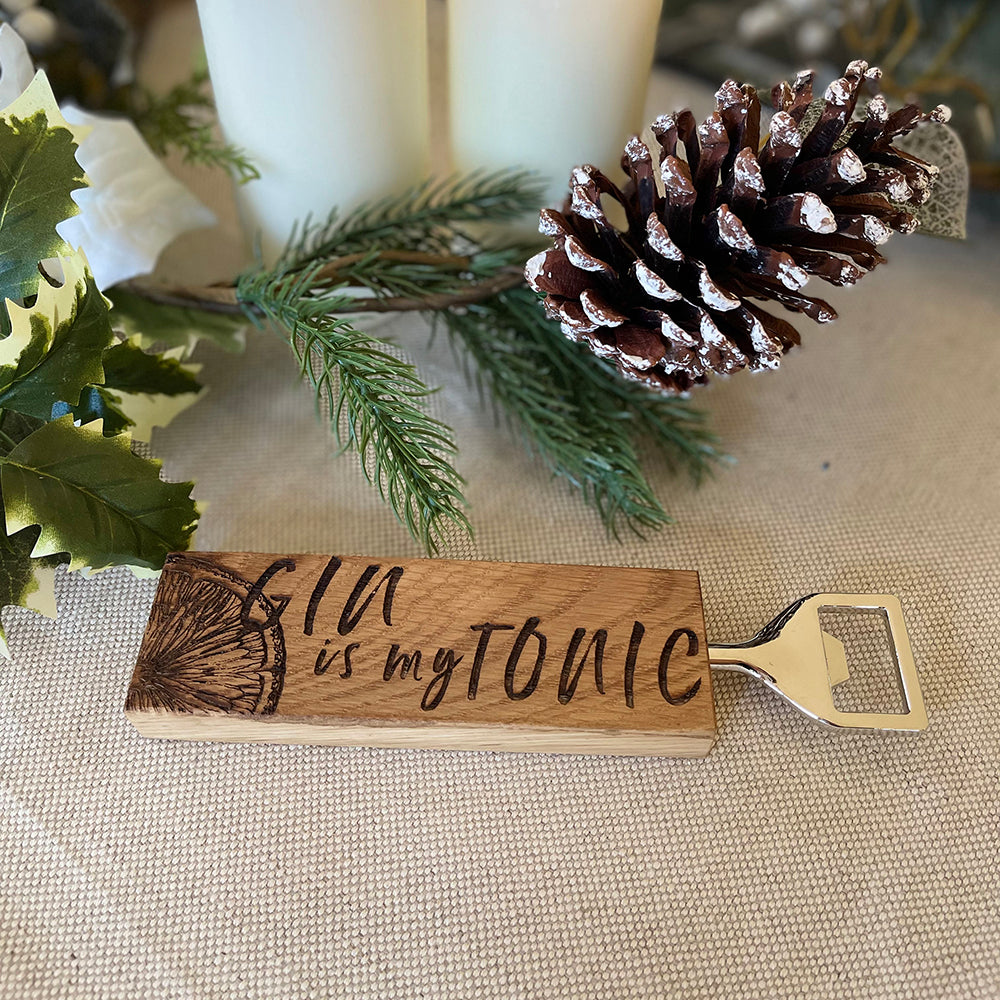 Ein Eichen Flaschenöffner mit einem eingebrannten Limonetten-Design und der Schrift "GIN is my TONIC" auf einem Tisch vor Zweigen und einem Tannenzapfen