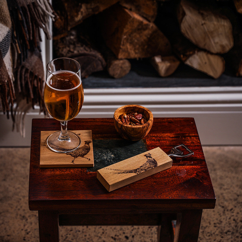 Ein Flaschenöffner mit einem rechteckigen Eichenholzgriff, auf dem ein Fasan-Motiv eingebrannt ist, dargestellt mit einer Schale Nüsse und einem Glas Bier