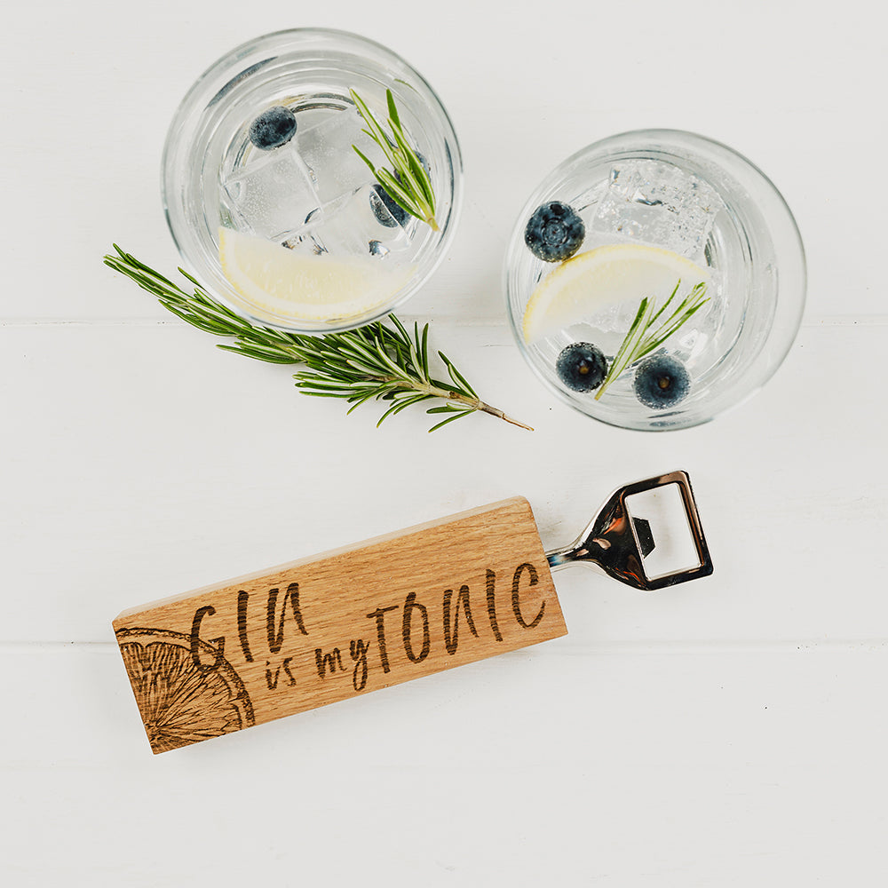 Ein Eichen Flaschenöffner mit einem eingebrannten Limonetten-Design und der Schrift "GIN is my TONIC", und zwei Gin Gläser in denen Eiswürfel, Zitronen und Blaubeeren enthalten sind