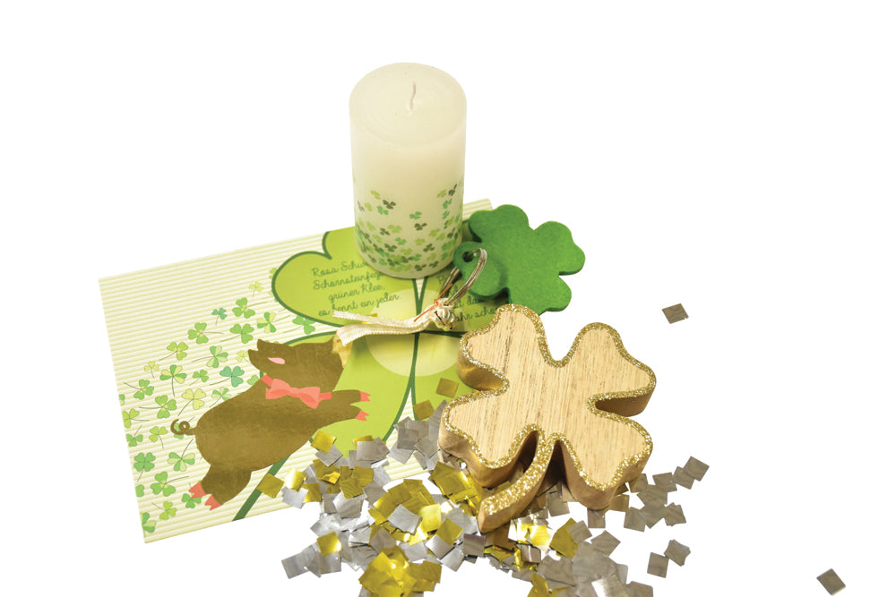 Postkarte mit einem Glücksschwein-Aufdruck, ein Holz Kleeblatt mit Goldumrandung, ein grünes Filz-Kleeblatt als Schlüsselanhänger, eine Kerze mit Kleeblatt-Muster und goldenes und silbriges Konfetti