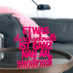 Wanddekoration Pinker Schriftzug "Etwas durchgeknallt ist ganz nah an wundervoll" auf einem Glastisch im Wohnzimmer