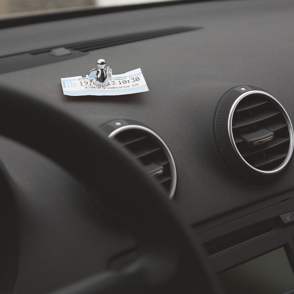 Angelo Schutzengel Parkscheinhalter auf dem schwarzen Ammaturenbrett im Auto mit einem Parkschein der zwischen Schutzengel und Podest geklemmt ist