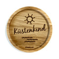 Eingekerbter Holzuntersetzer mit dem aufgedruckten Spruch „Küstenkind" mit aufgedruckter Sonne und Wasser-Linien