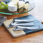 Drei silberne glänzende Käse Serviermesser aus Edelstahl auf einem Holzbrett angerichtet mit Käseplatte im Hintergund