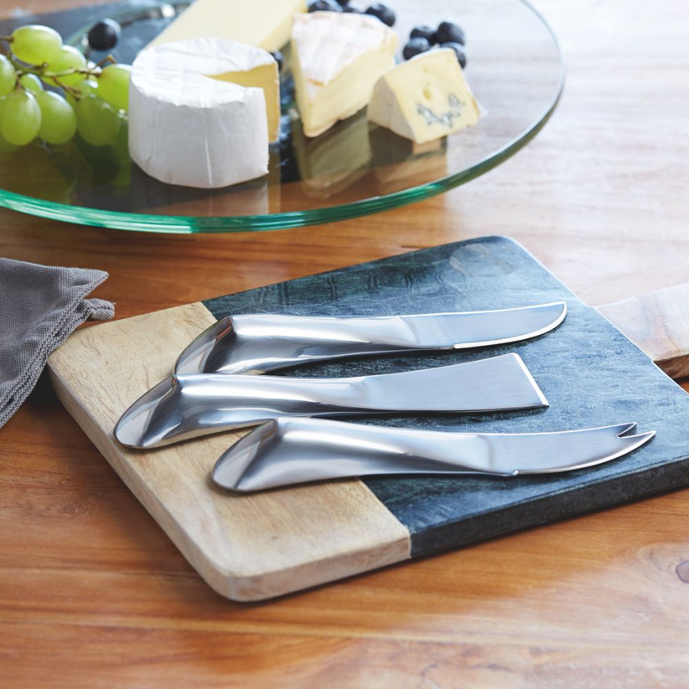 Drei silberne glänzende Käse Serviermesser aus Edelstahl auf einem Holzbrett angerichtet mit Käseplatte im Hintergund