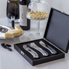 Drei silberne glänzende Käse Serviermesser in einer edlen schwarzen Holzschatulle mit Polsterung und Metallverschluss angerichtet mit Käseplatte und Wein im Hintergund