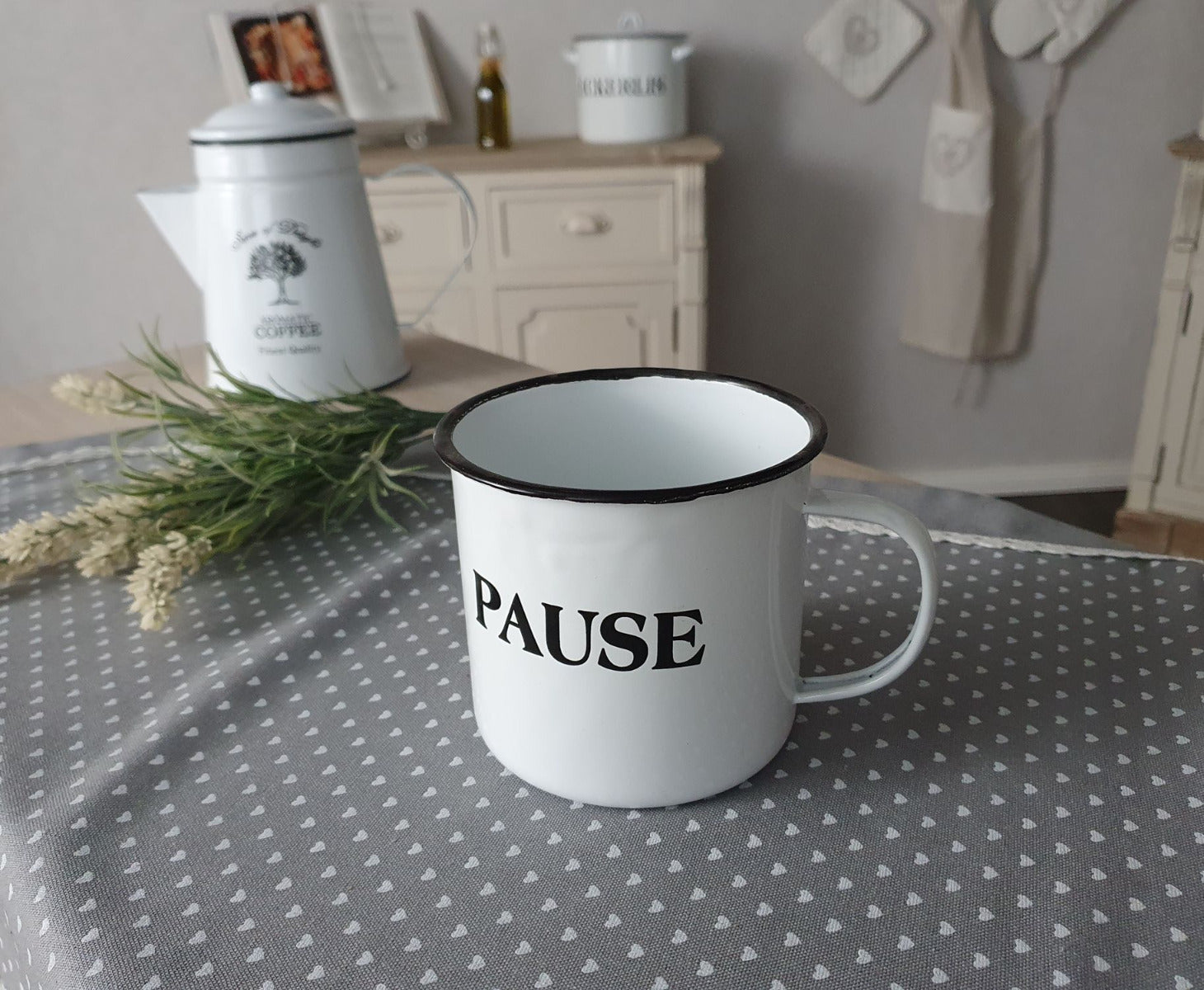 Weißer Becher mit der schwarzen Schrift "PAUSE" auf einem Tisch mit einer Kaffekanne im Hintergrund