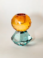 Ein kleiner Kerzenhalter aus Glas in Petrol und Bernstein Kristall-Optik, bestehend aus mehreren geometrischen Flächen. Der Kerzenhalter hat eine moderne, facettierte Optik, die das Licht reflektiert.