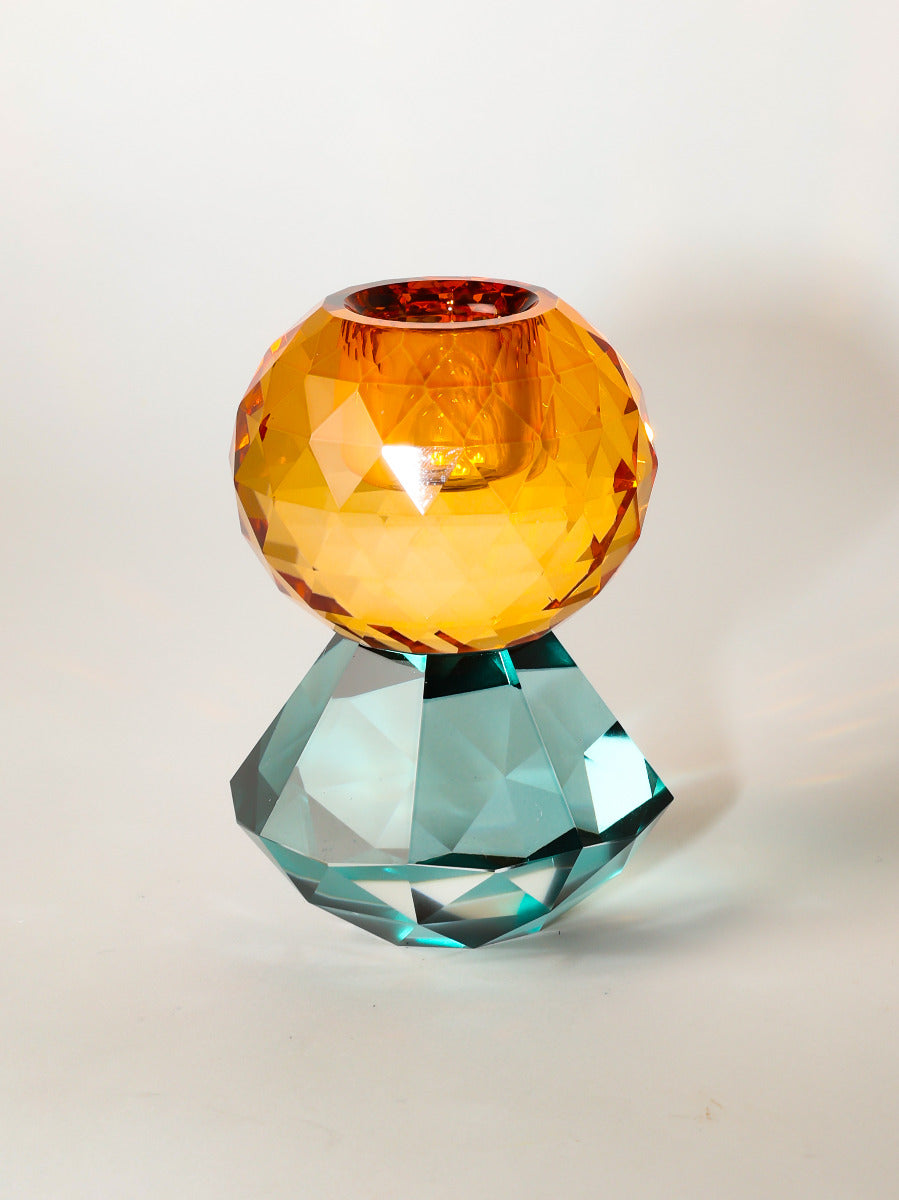 Ein kleiner Kerzenhalter aus Glas in Petrol und Bernstein Kristall-Optik, bestehend aus mehreren geometrischen Flächen. Der Kerzenhalter hat eine moderne, facettierte Optik, die das Licht reflektiert.