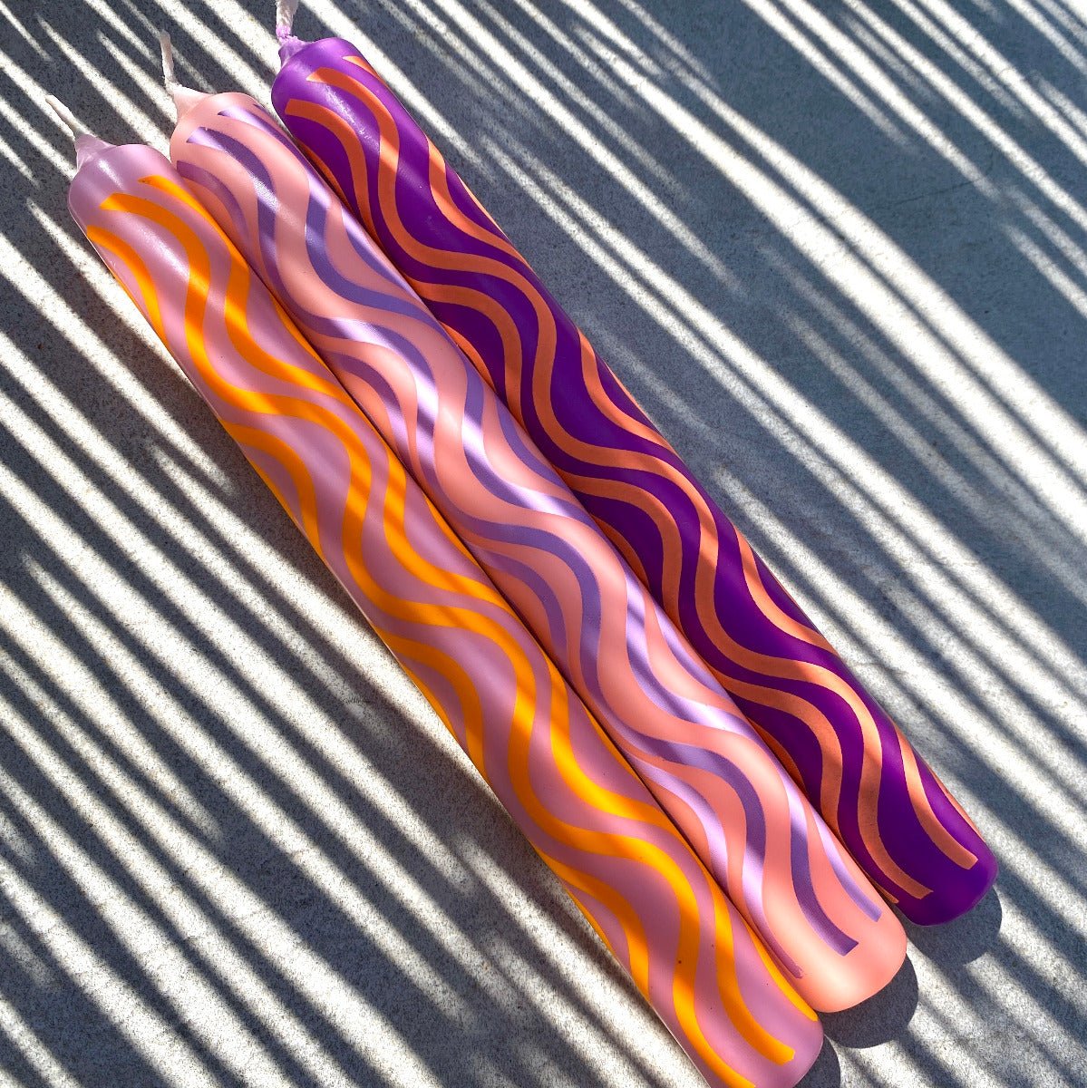 Drei Stabkerzen mit Wellenmusterung in den Farben Lila, Pink, und Orange, in der Sonne