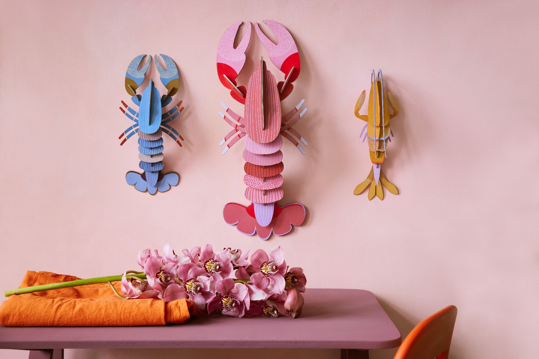 Drei verschiedenfarbige Hummer Wanddekorationen aufgehangen an einer Pinken Wand über einem Tisch