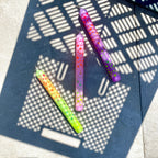 Drei Stabkerzen in regenbogen Farben mit Punkten, die ebenfalls in vielen Farben auf der Kerze sind, liegen in teils Sonne und Schatten