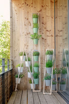 Drei Pflanzleiter mit blassgrünen Kübeln im Milcheimer Design. Eines ist an der Wand angelehnt und zwei andere sind ausgebreitet als Stehleiter und enthalten verschiedene Pflanzen auf dem Balkon