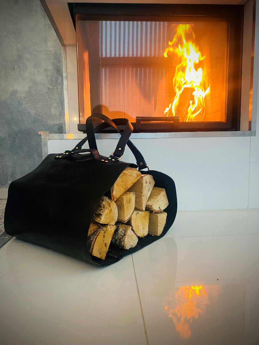 Brennholztragetasche aus Leder in schwarz gefüllt mit Brennholz für den Kamin, Kamin mit Feuer im Hintergrund, gemütliche Atmosphäre, Holzträger liegt auf dem Boden vor dem Kamin