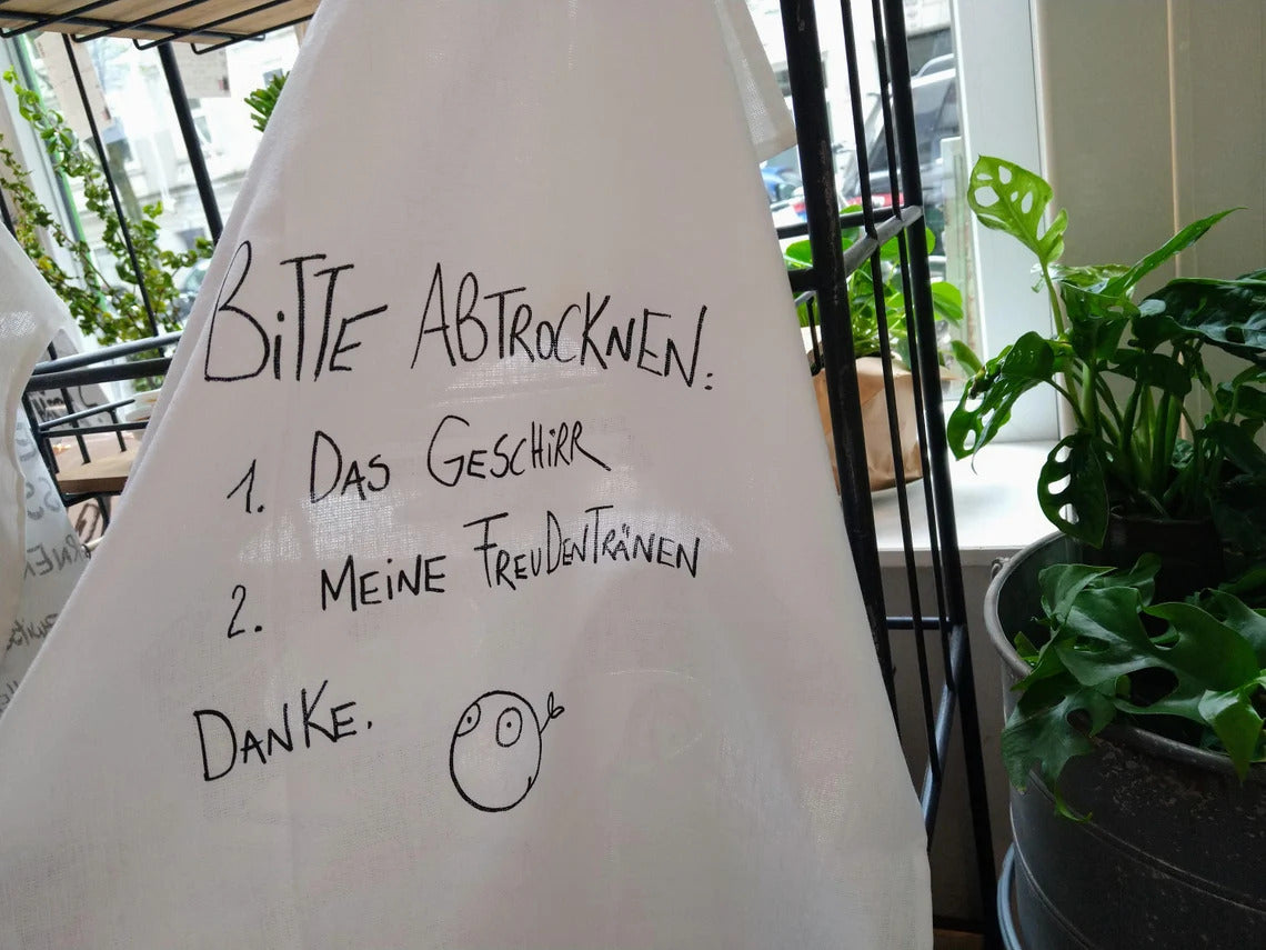 Aufgehangenes Geschirrtuch mit der schwarzen Schrift "Bitte Abtrockenen: 1.Das Geschirr  2.Meine Freudentränen Danke."