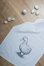 weißes Geschirrtuch aus Baumwolle mit einer aufgedruckten schwarzen Ente auf Laminatboden ausgelegt auf dem auch eier und Knoblauch liegen