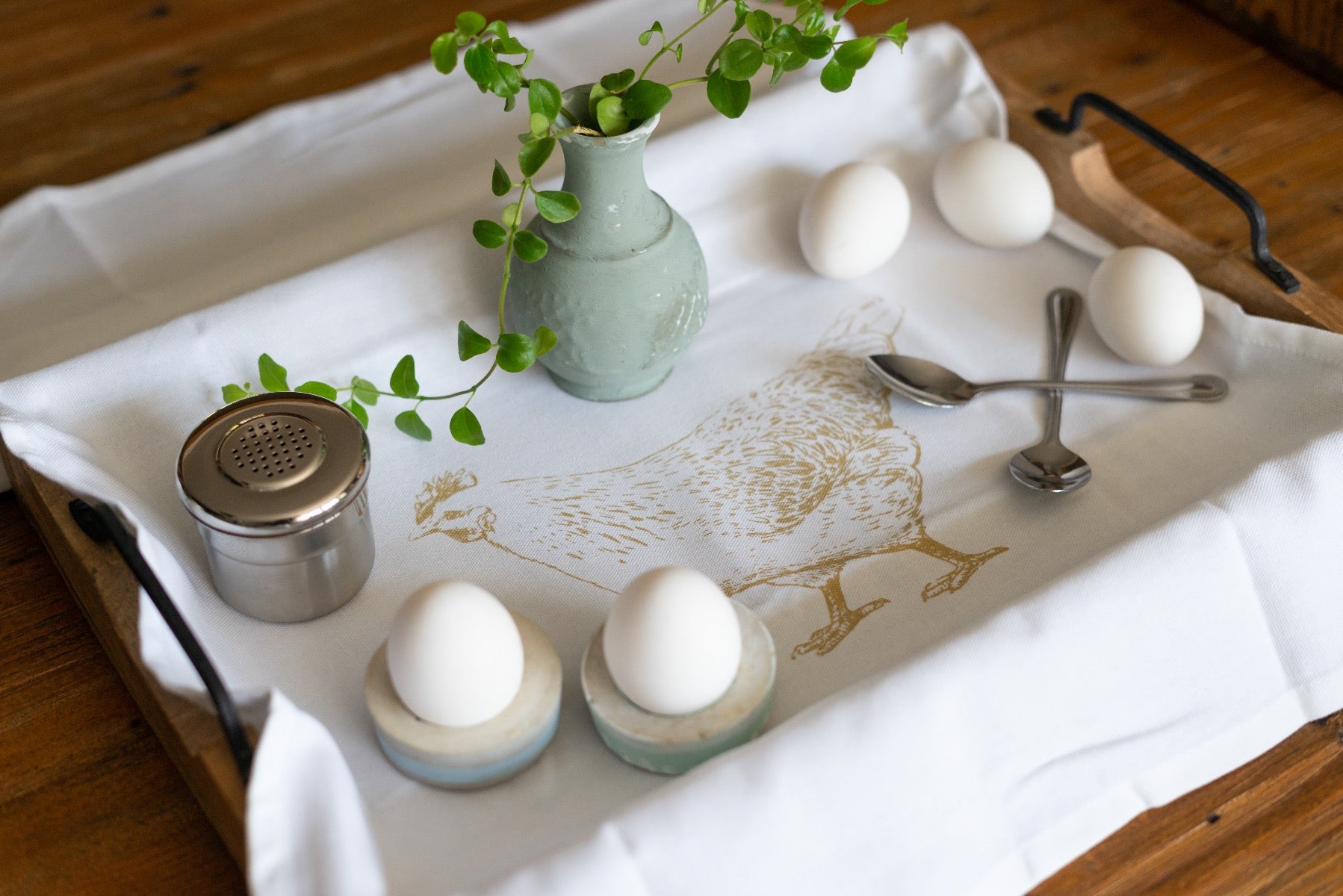 Das Baumwoll-Geschirrtuch mit goldfarbenem Huhn-Aufdruck zusammen auf einem Tablett mit Hühnereier, Besteck und Eierhalter