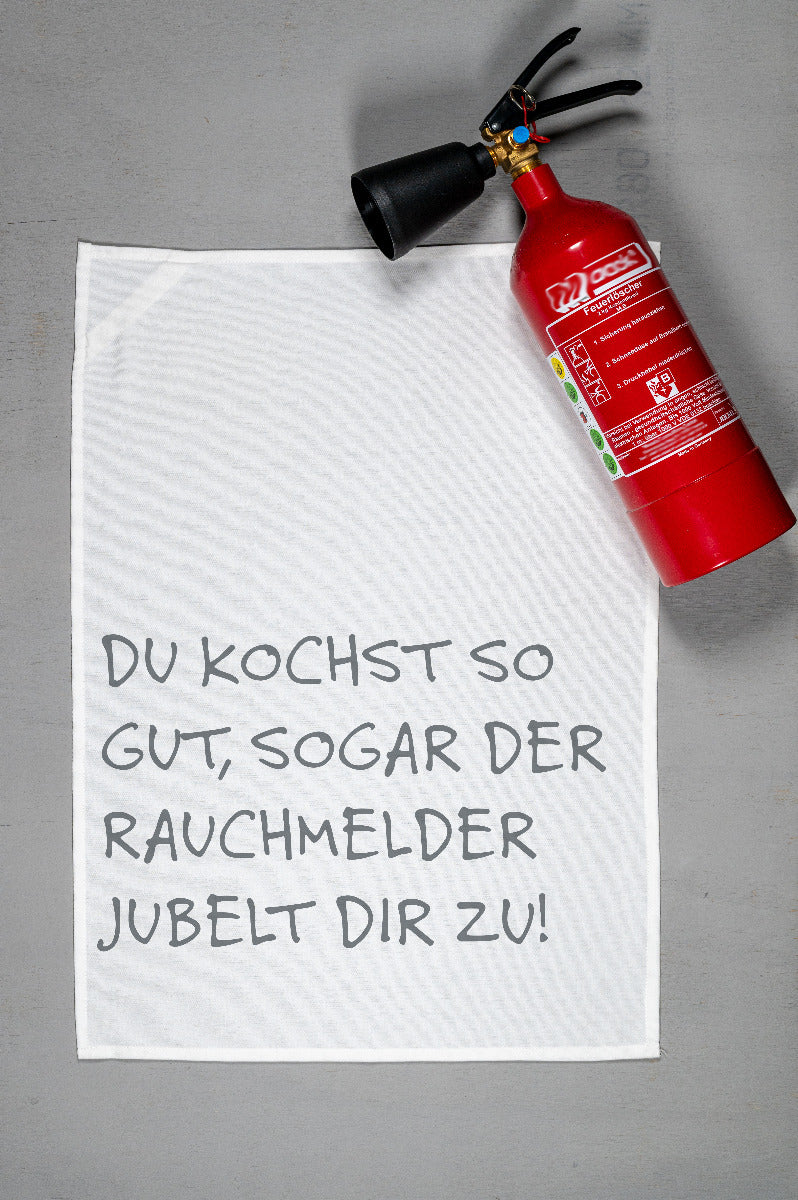 Weißes Geschirrtuch aus Baumwolle liegt auf dem Küchentresen und daneben liegt ein roter kleiner Rauchmelder