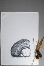 Weißes Geschirrtuch mit grauer Illustration eines Igels, der sich entspannt an einen Apfel lehnt mit Weizen und Tannenzapfen daneben