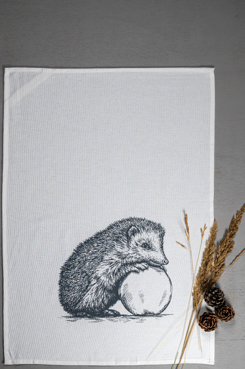 Weißes Geschirrtuch mit grauer Illustration eines Igels, der sich entspannt an einen Apfel lehnt mit Weizen und Tannenzapfen daneben