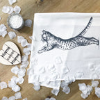 Das Geschirrtuch mit Katzen-Motiv auf dem Tisch mit weißen Blütenblättern und einem Holzherz mit der Schrift "Home Sweet Home"