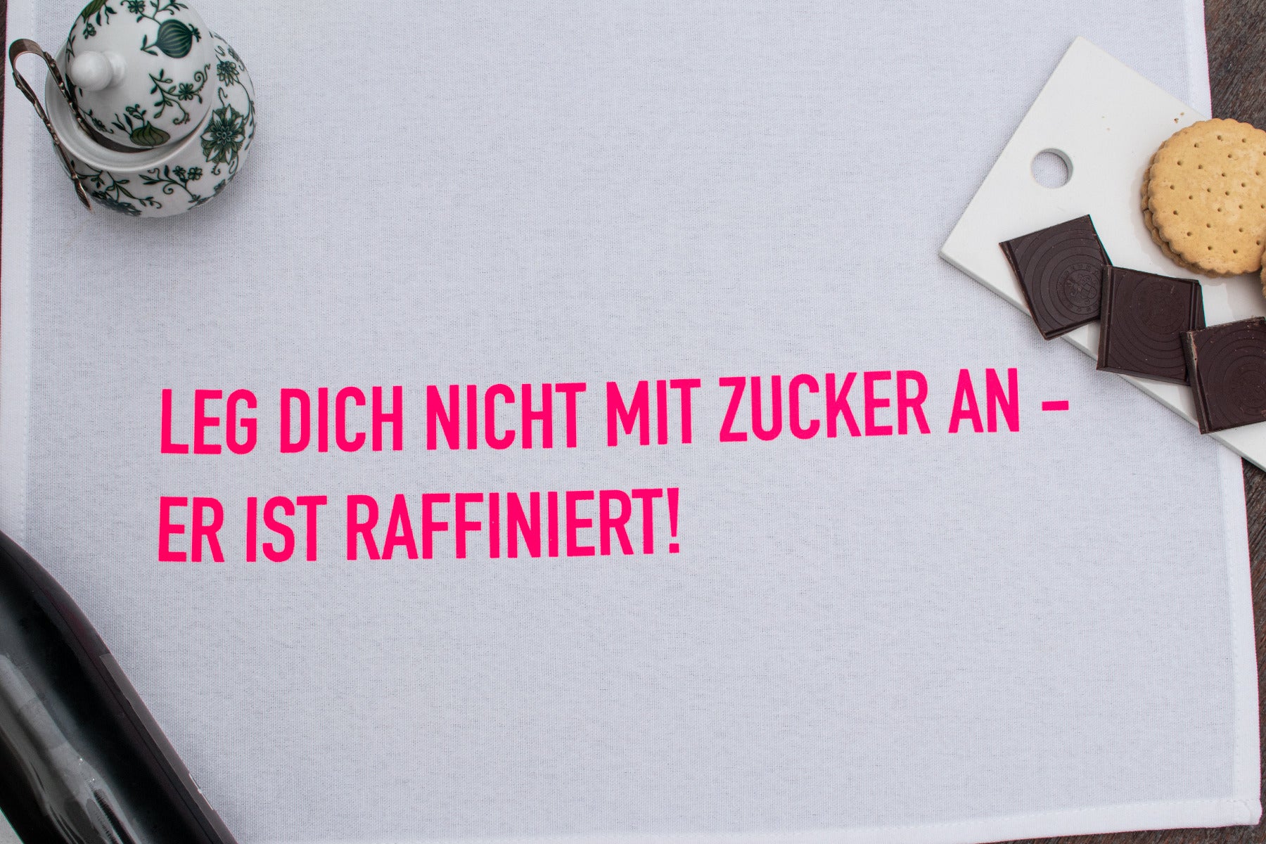 Das Geschirrtuch mit der Schrift "Leg dich nicht mit zucker an - Er ist raffiniert" auf dem Küchentisch mit Plätzchen 
