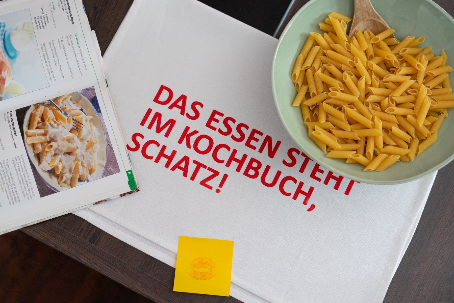 Weißes Geschirrtuch mit einer Roten Schrift "DAS ESSEN STEHT IM KOCHBUCH, SCHATZ!" auf dem Tisch ausgebreitet auf dem eine Schüssel Nudel mit aufgeklapptem Kochbuch steht