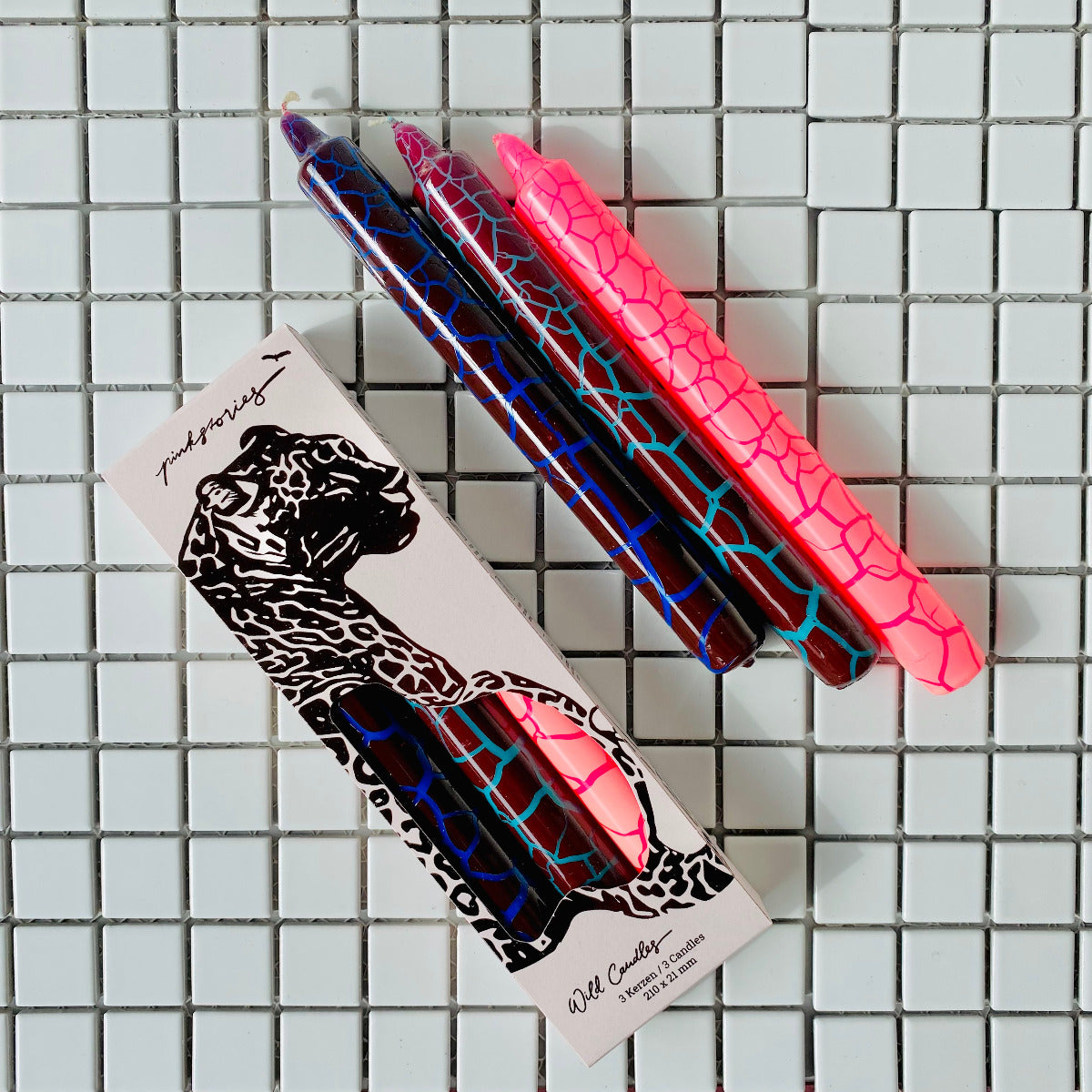 Drei Stabkerzen in Pink, Blau, Hellblau mit einem Trockenriss Muster und eingepackte auf einem Fliesenteppich