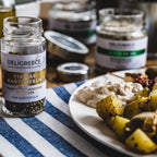 Fläschchen Wildkräutermischung "YIAYIAS Kartoffeln" von Deligreece auf einem gedeckten Tisch mit zubereitetem Essen