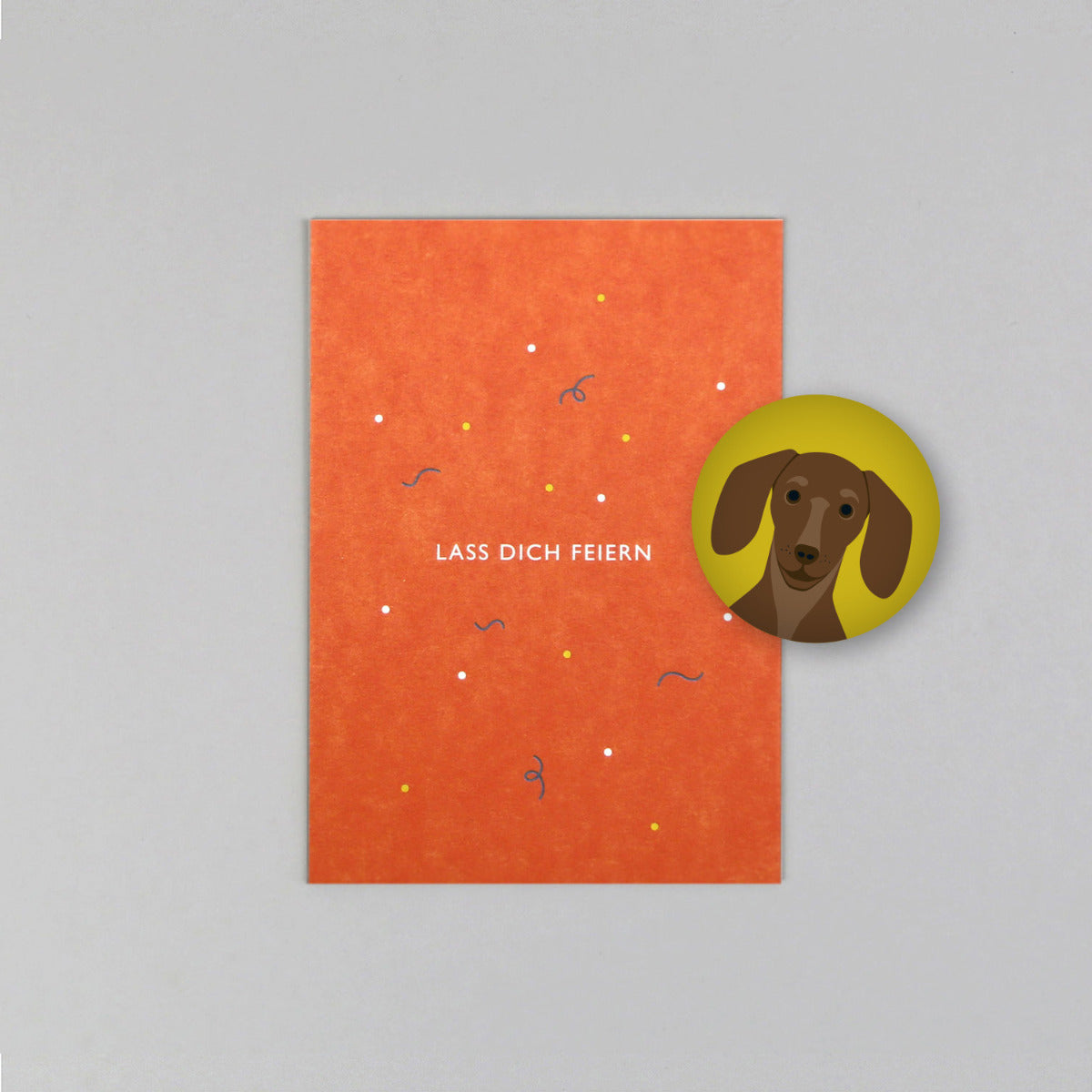 Orangene Postkarte mit Schrift "Lass dich feiern" mit gelbem Magnet mit Dackel Motiv Grauer Hintergrund